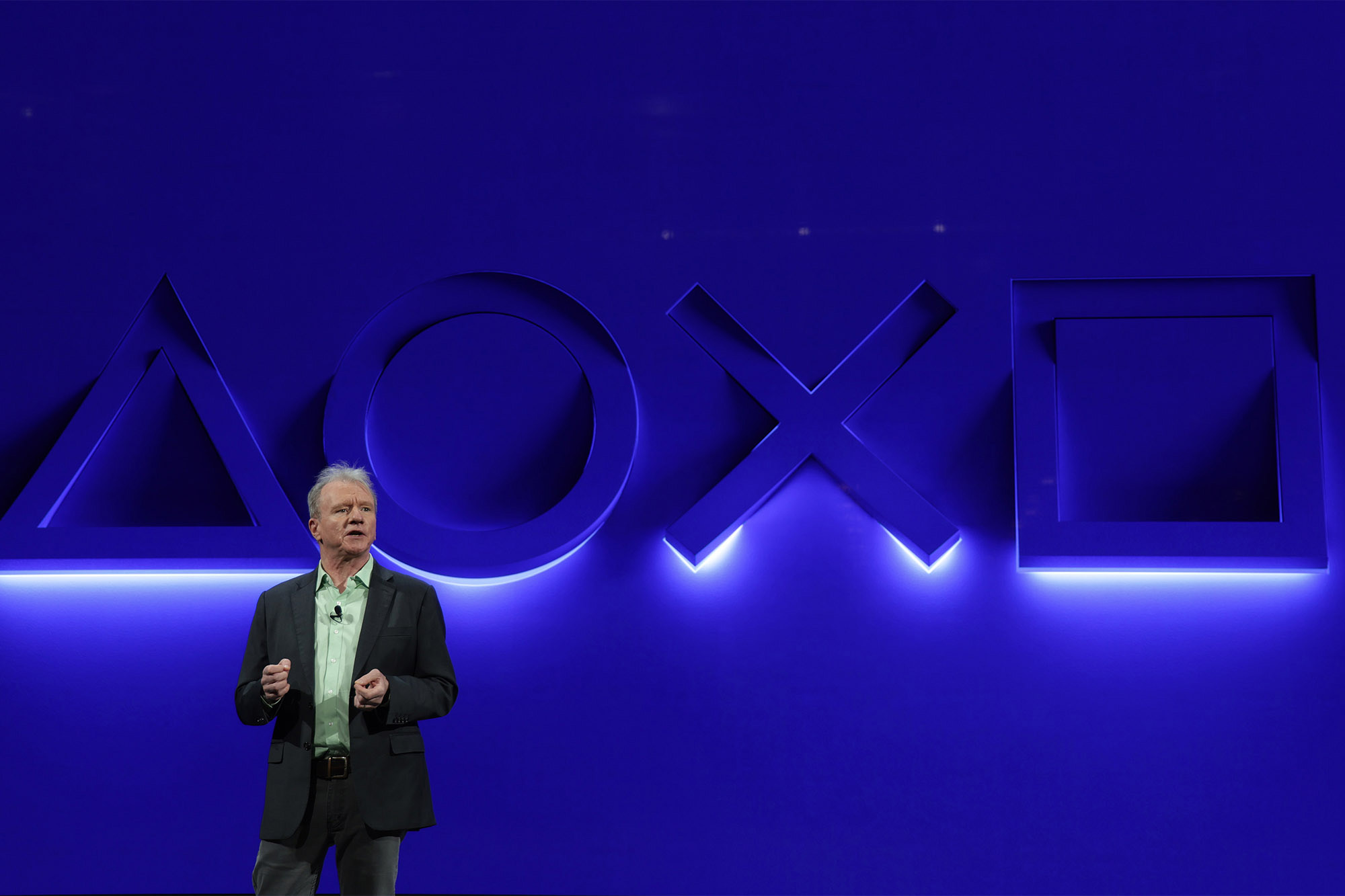 Playstation CEO and president, Jim Ryan, stands in front of a blue wall with Playstation button symbols illuminated.