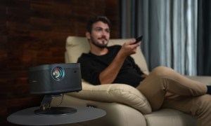 A man in a chair with a remote, and a projector on a table.