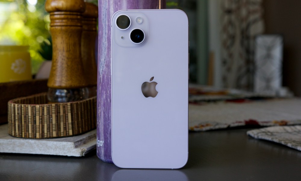 An iPhone 14 standing up on a table. Behind it is a purple water bottle.