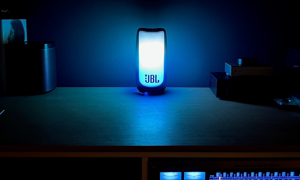 The JBL Pulse 5 Bluetooth speaker, glowing blue on a media sideboard table with a stereo receiver.