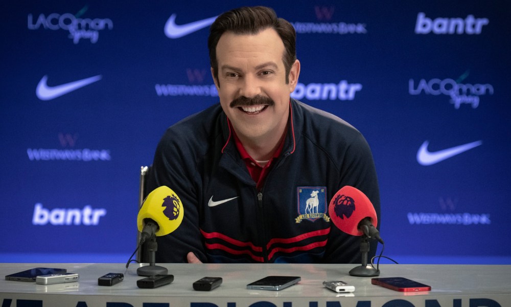 Ted Lasso sits at a press conference table in season 3.