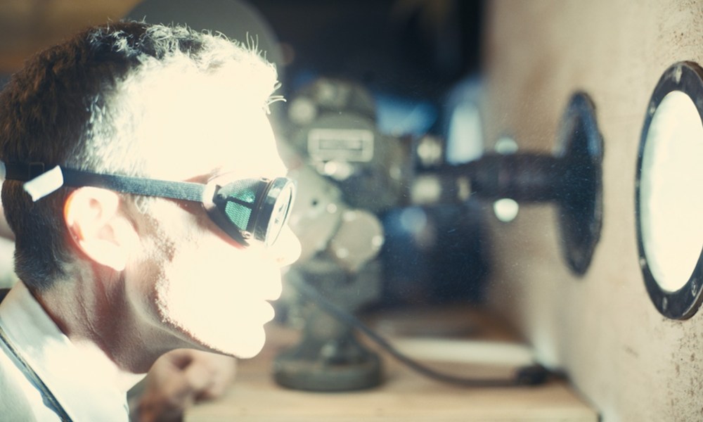 Cillian Murphy stares through a small window at an atomic blast, his face illuminated by light.