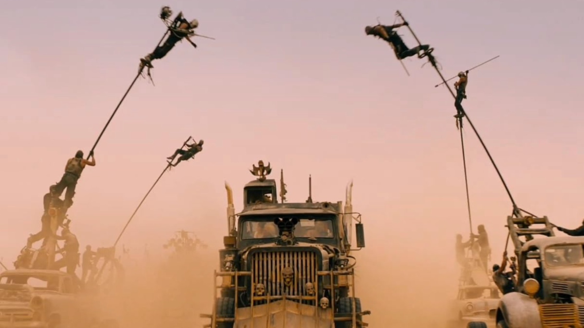 Men swing from poles while driving cars in the desert.