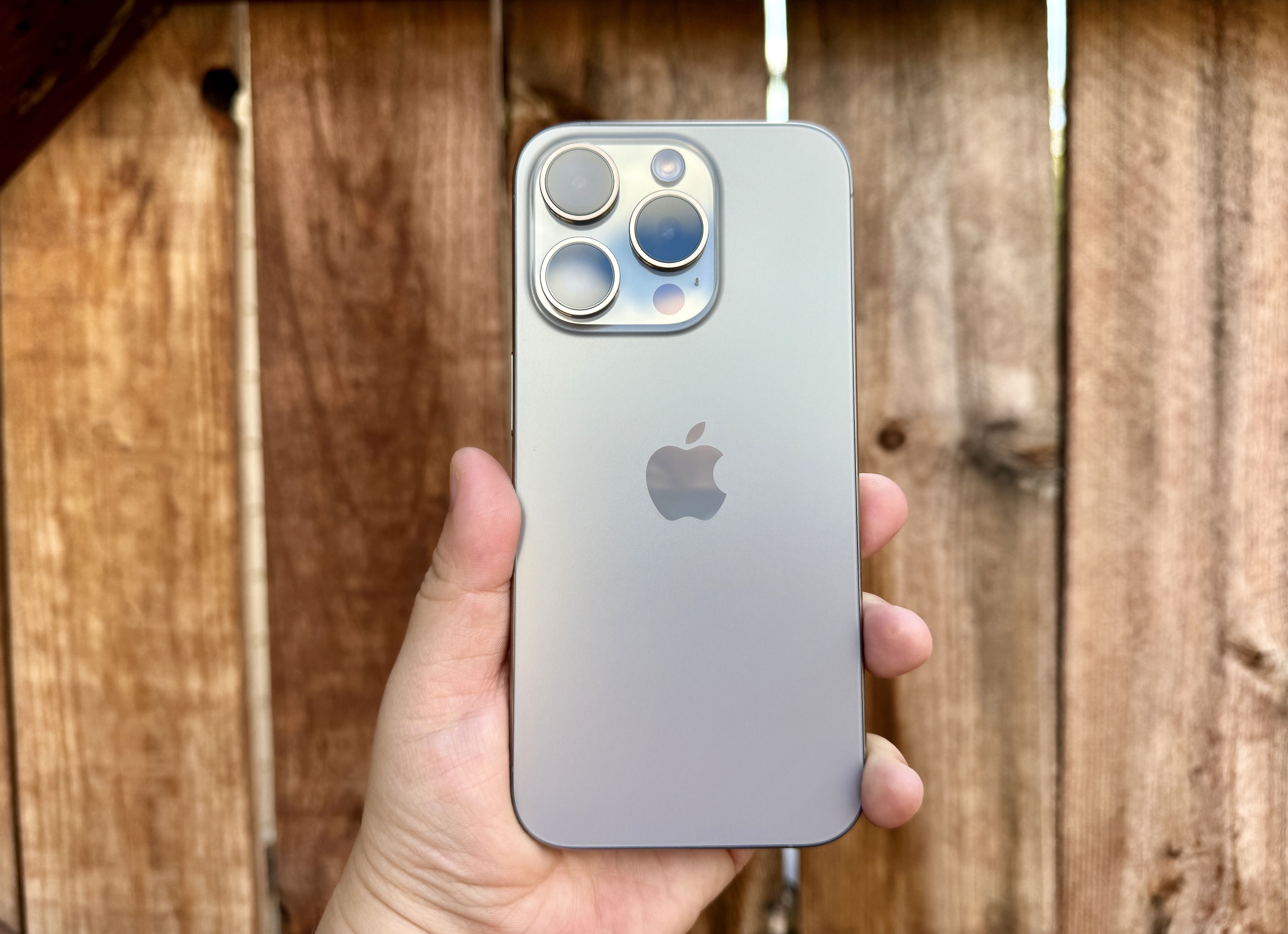 iPhone 15 Pro in Natural Titanium held in hand in front of a wooden gate.