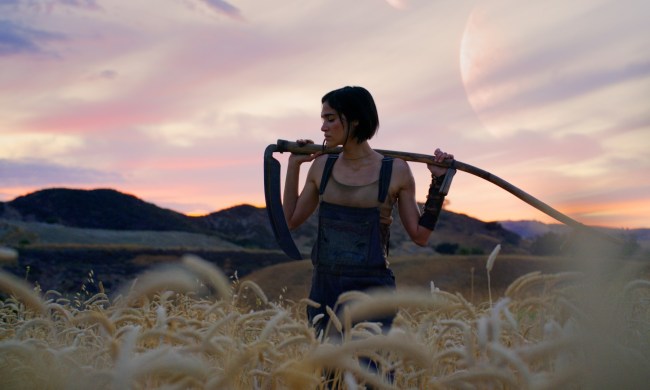 Sofia Boutella strikes a cool pose with a stick against a pretty landscape in a still from Rebel Moon – Part One: A Child of Fire.