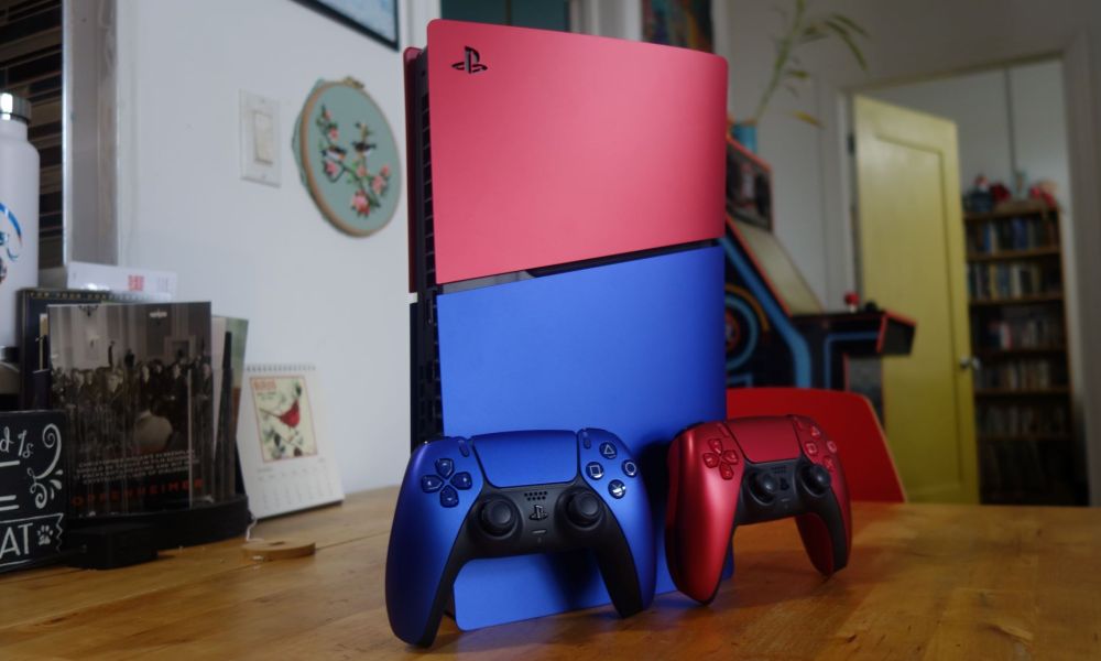 A red and blue PS5 stands on a table with matching controllers.