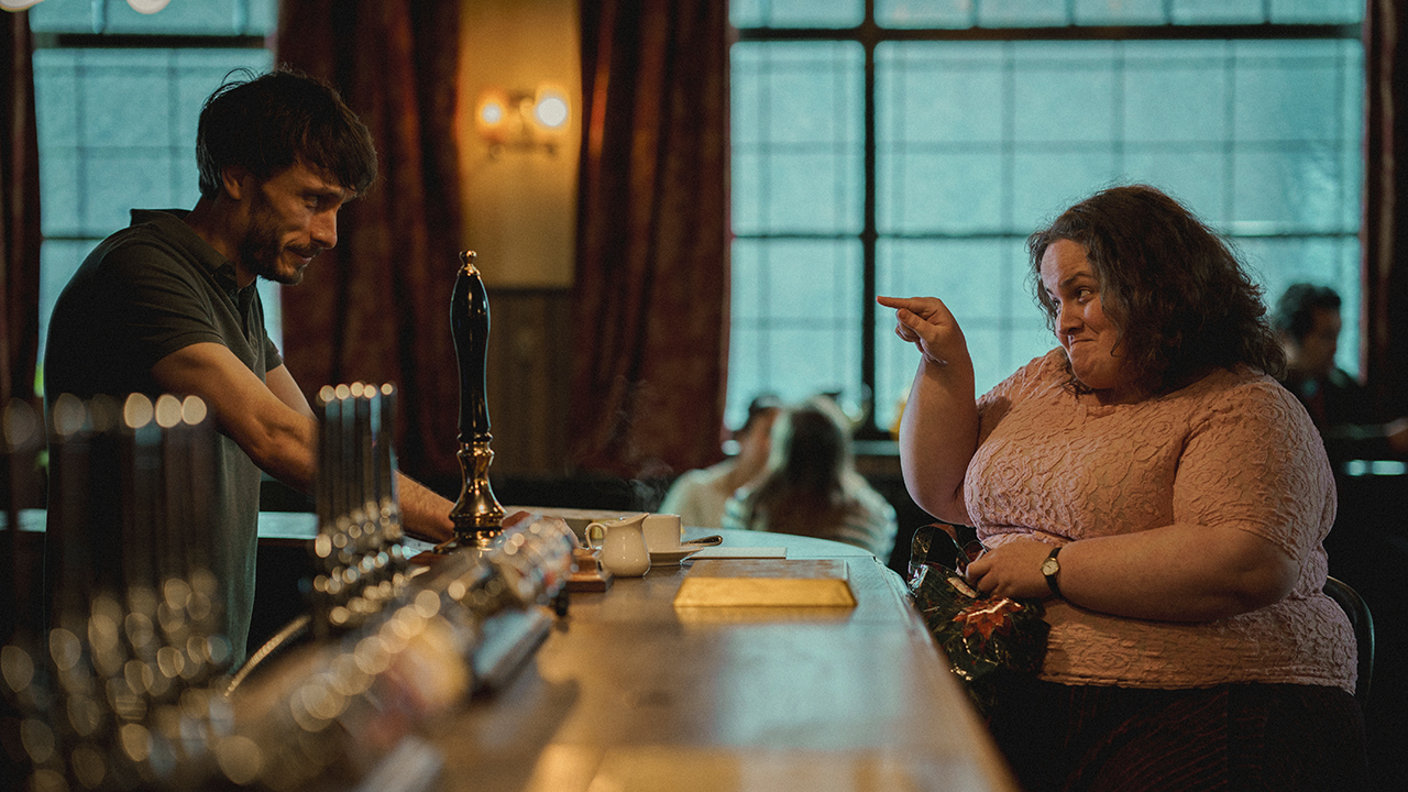 A man behind a bar, a heavyset woman sitting there and pointing at him, both smiling in a scene from Baby Reindeer.