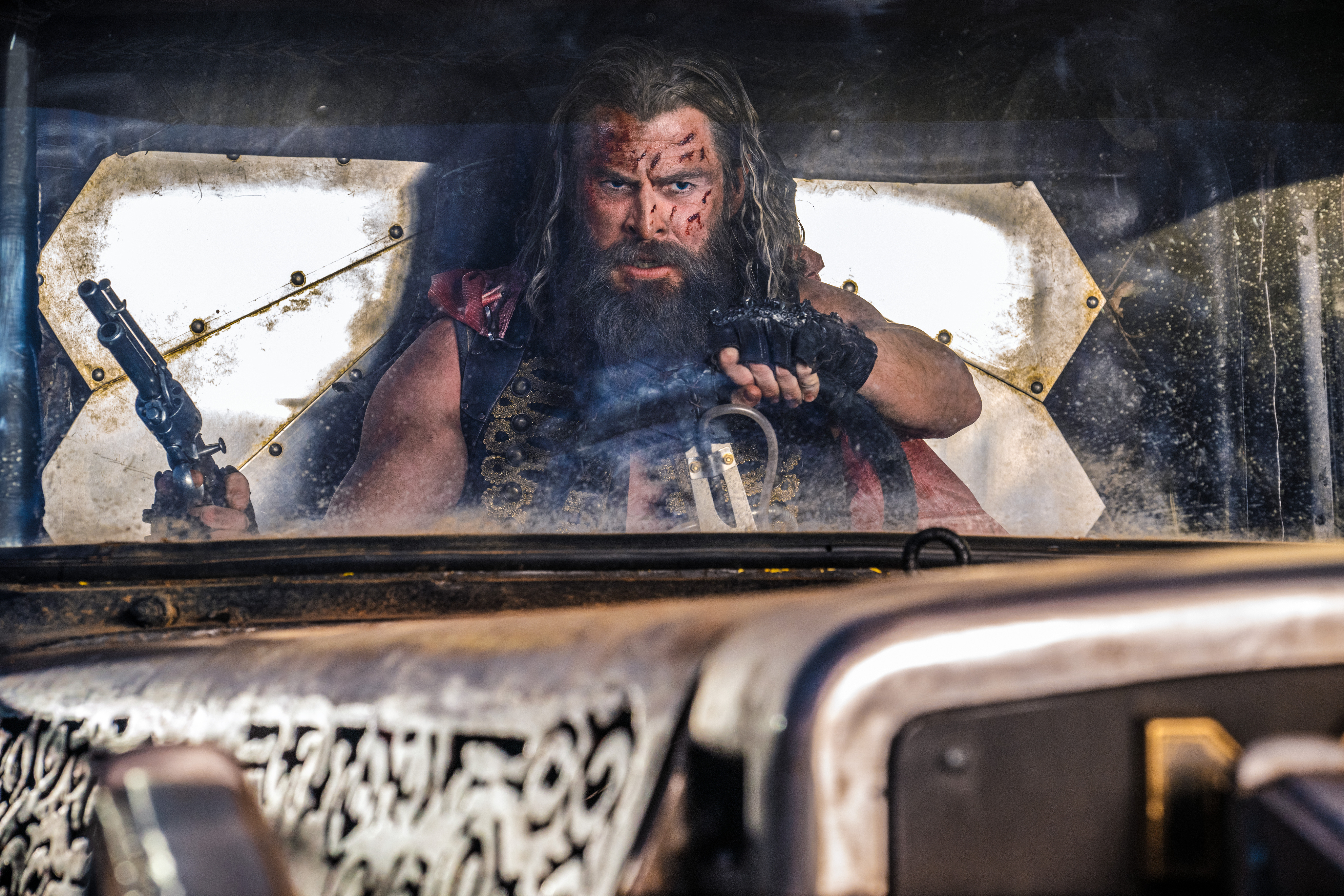 A bloodied Chris Hemsworth holds a gun and a steering wheel in Furiosa: A Mad Max Saga.