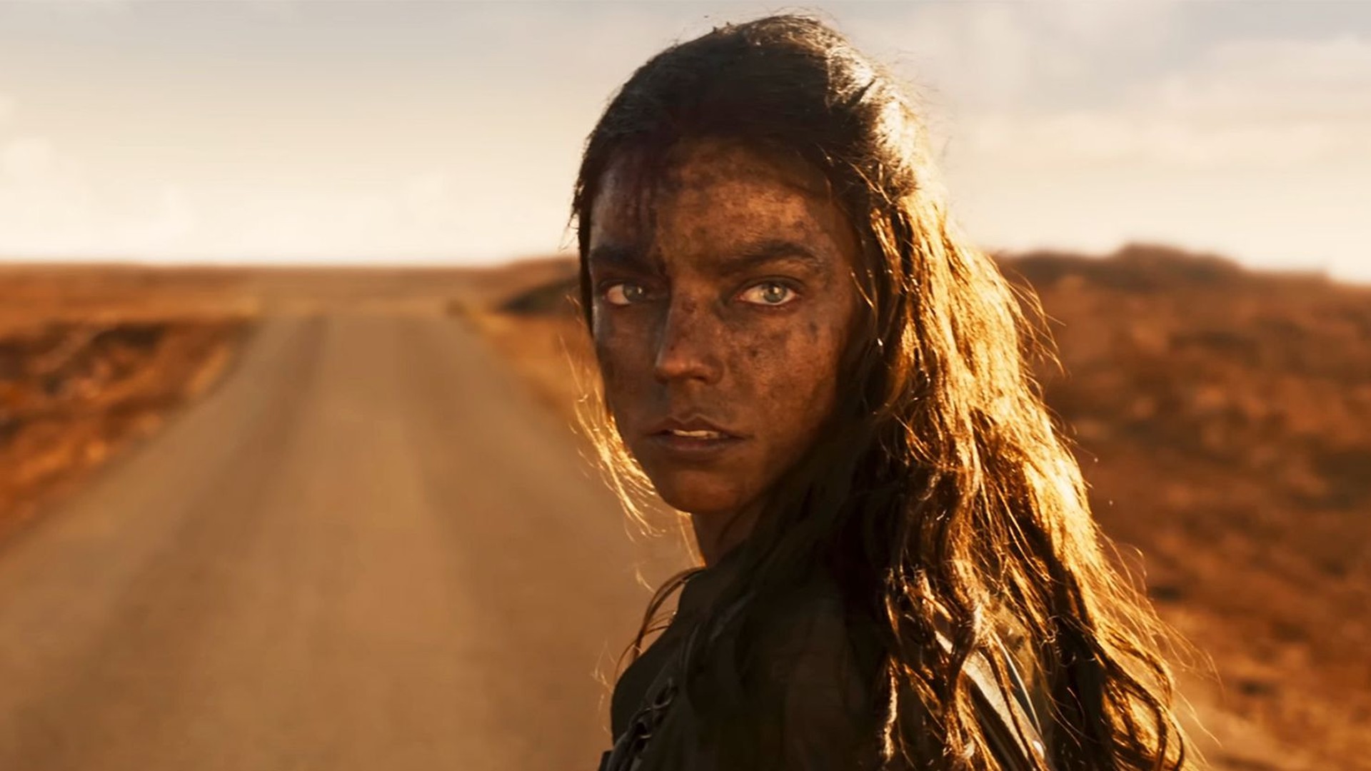 Anya Taylor-Joy stands on a dusty road and glances over her shoulder in a still from Furiosa: A Mad Max Saga.