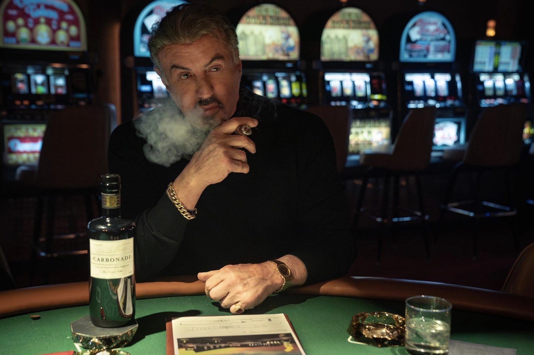 Sylvester Stallone smokes a cigar while sitting at a table in Tulsa King.