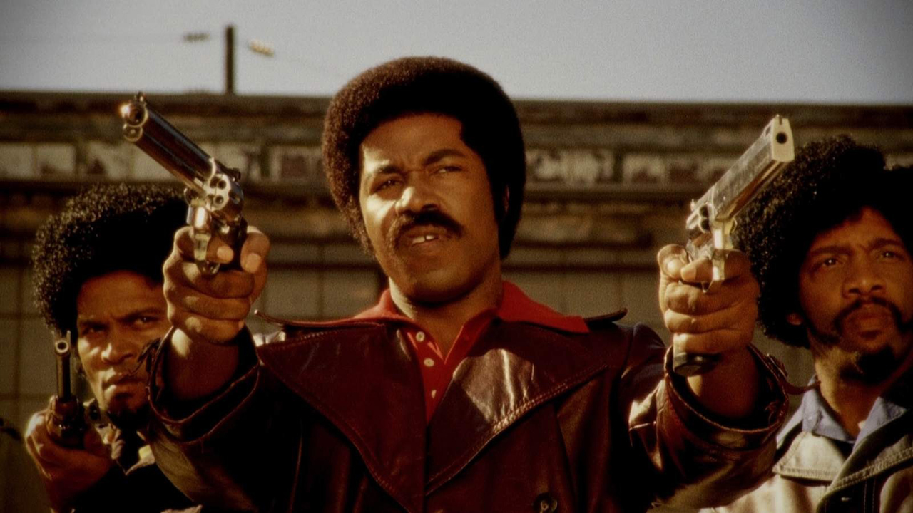 A man holds up two guns with two others behind him in a scene from Black Dynamite.