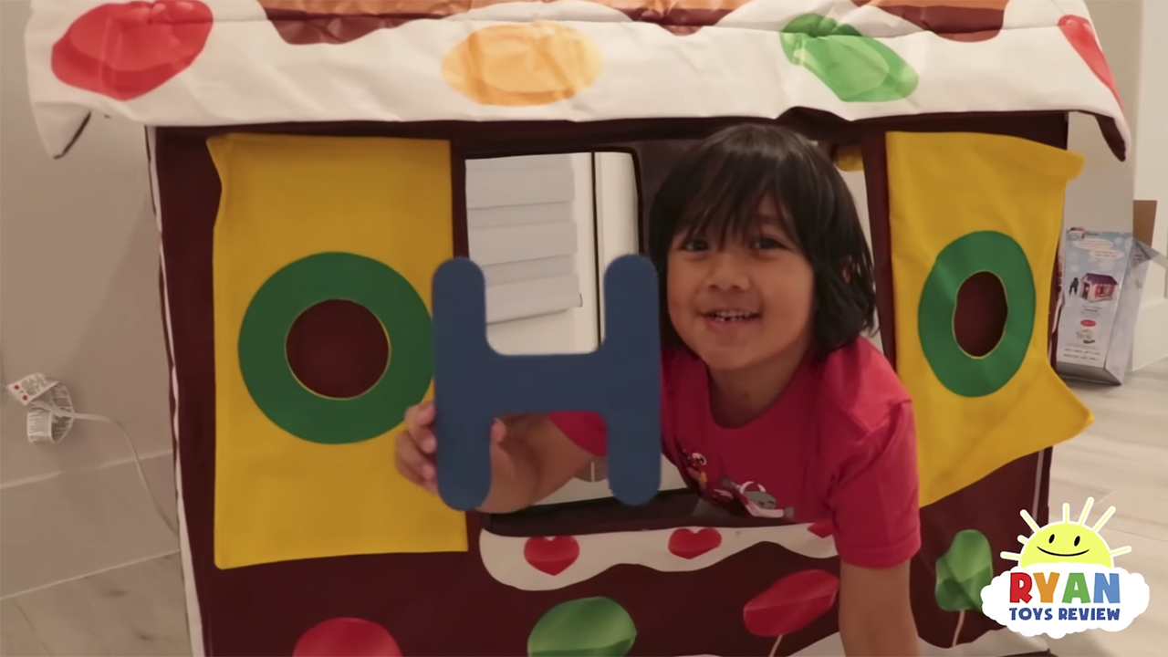 Ryan from Ryan's World holding up a letter H in front of a playhouse and smiling.