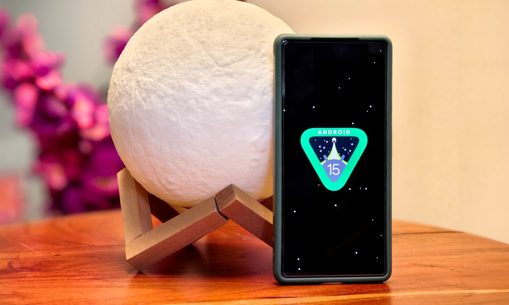 Android 15 easter egg shown on a Google Pixel 6a kept on a table in front of moon shaped lamp and pink flowers.