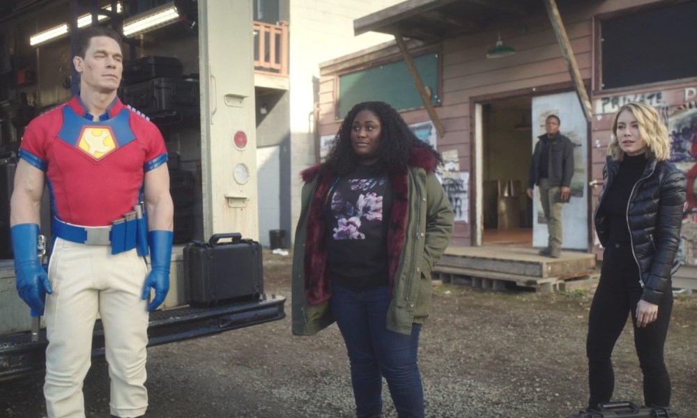 Peacemaker stands next to two women and stare.