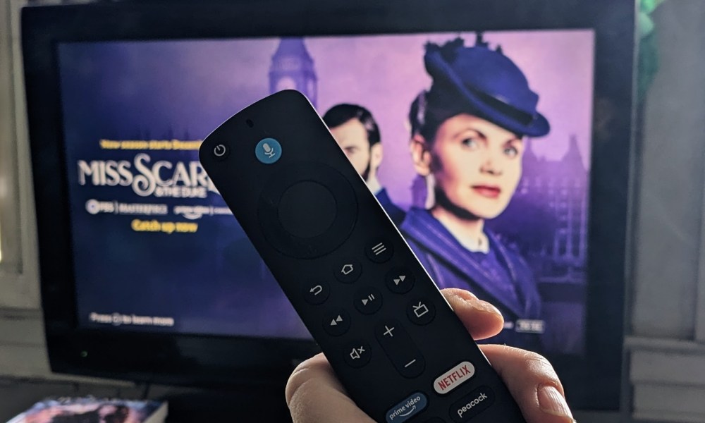 An Amazon Fire TV remote is held up in front of a TV. In the background a show is obscured by the remote.