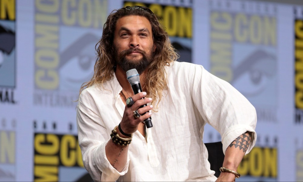 Jason Momoa holds a mic while sitting down and staring.