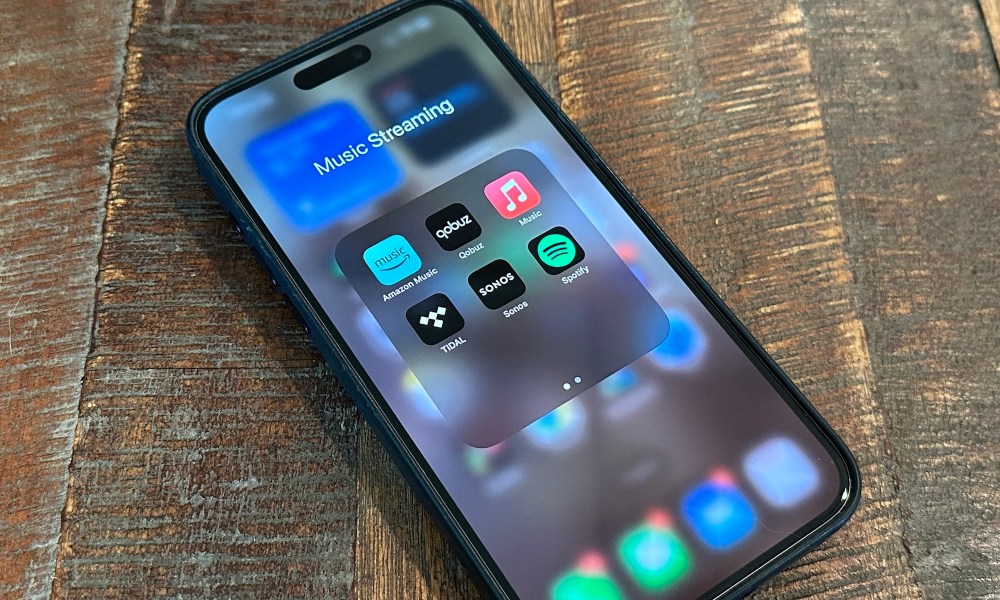 An iPhone 15 Pro Max on a wooden table displaying streaming apps Amazon Music, Qobuz, Apple Music, Tidal, Sonos, and Spotify.