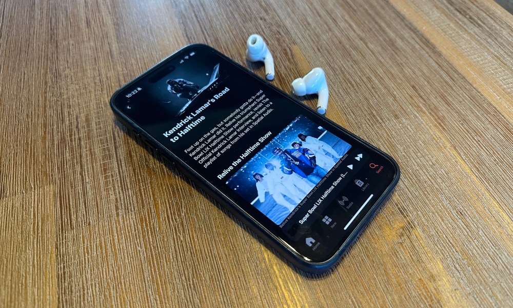 An iPhone 15 Max displaying the Super Bowl LIX Halftime Show with Apple AirPods Pro 2 next to it on a wooden table.